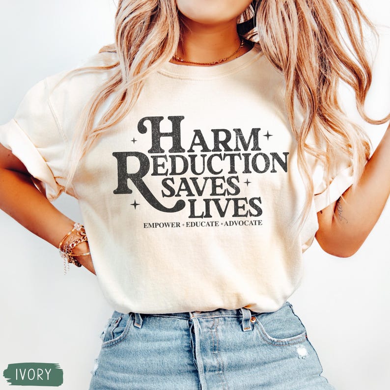 May include: Ivory-colored t-shirt with the text "Harm Reduction Saves Lives" in a bold, black font. The shirt also includes the words "Empower. Educate. Advocate." in a smaller font. The model is wearing blue jeans.