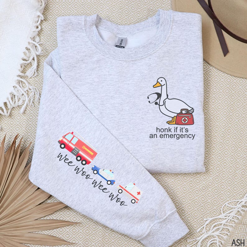 May include: Light gray sweatshirt with a cartoon duck wearing a stethoscope and carrying a medical bag. The text "honk if it's an emergency" is below the duck. The left sleeve has a fire truck, police car, and ambulance with the text "wee woo wee woo".