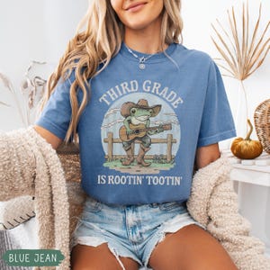May include: Blue jean colored t-shirt with the text "THIRD GRADE IS ROOTIN' TOOTIN'". The graphic features a cartoon frog wearing a cowboy hat and boots, playing a guitar in front of a wooden fence.