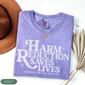May include: A violet Comfort Colors t-shirt with the text "Harm Reduction Saves Lives" in white. The shirt has a crew neck and short sleeves. A gold necklace with a pendant is draped on the shirt. A brown hat and a dried palm leaf are in the background.