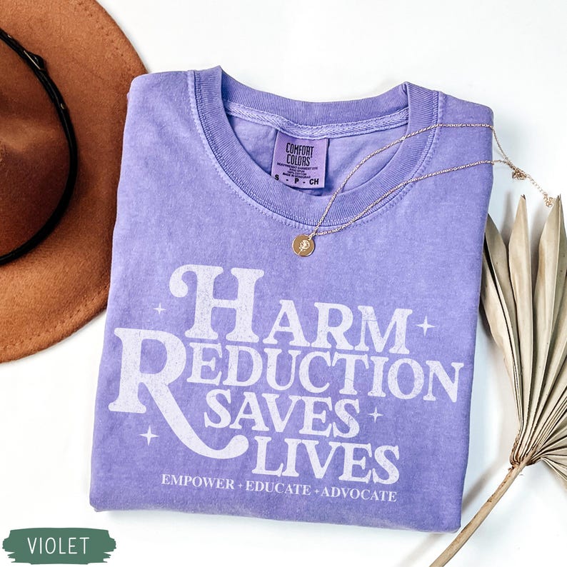 May include: A violet Comfort Colors t-shirt with the text "Harm Reduction Saves Lives" in white. The shirt has a crew neck and short sleeves. A gold necklace with a pendant is draped on the shirt. A brown hat and a dried palm leaf are in the background.