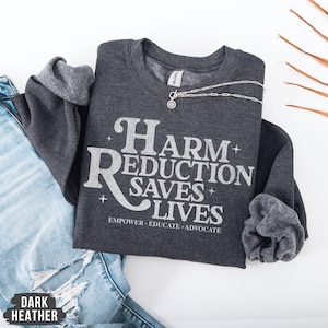 May include: Dark heather grey sweatshirt featuring the text "Harm Reduction Saves Lives" in white. The sweatshirt has a crew neck and long sleeves. A silver necklace with a pendant is draped across the front. The words "Empower, Educate, Advocate" are printed below the main text.