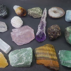 May include: A collection of various crystals and stones, including amethyst, rose quartz, clear quartz, selenite, tiger's eye, emerald, and agate. The crystals are arranged on a dark surface.