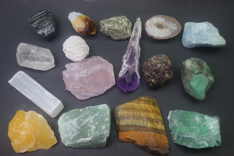 May include: A collection of various crystals and stones, including amethyst, rose quartz, clear quartz, selenite, tiger's eye, emerald, and agate. The crystals are arranged on a dark surface.