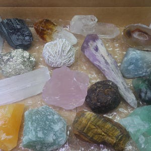 May include: A collection of various crystals and stones in a box. The crystals are in different shapes, sizes, and colors, including clear, white, pink, purple, green, brown, and yellow.