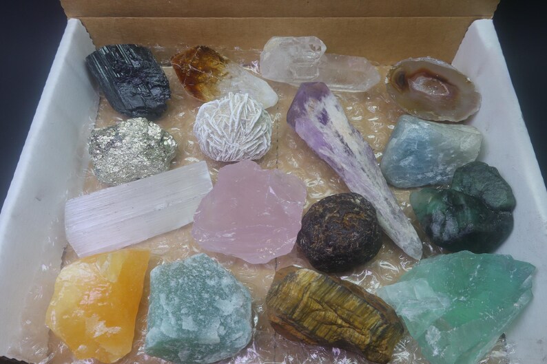 May include: A collection of various crystals and stones in a box. The crystals are in different shapes, sizes, and colors, including clear, white, pink, purple, green, brown, and yellow.