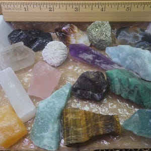 May include: A collection of 15 different types of crystals and stones in a white box. The crystals are various colors, including clear, white, pink, green, blue, purple, brown, and yellow. The stones are arranged in a random pattern.
