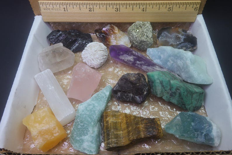 May include: A collection of 15 different types of crystals and stones in a white box. The crystals are various colors, including clear, white, pink, green, blue, purple, brown, and yellow. The stones are arranged in a random pattern.