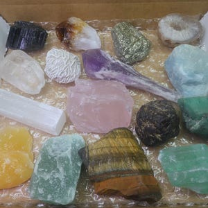 May include: A collection of 15 different types of crystals and stones in a cardboard box. The crystals are various colors, including white, pink, green, purple, brown, and yellow.