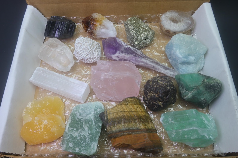 May include: A collection of 15 different types of crystals and stones in a cardboard box. The crystals are various colors, including white, pink, green, purple, brown, and yellow.