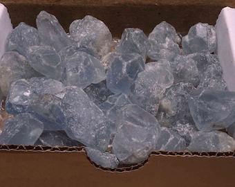 Puede incluir: Una caja llena de una gran cantidad de cristales de corte tosco translúcidos de color azul claro. Los cristales son de varios tamaños y formas, y parecen ser naturales.