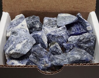 Puede incluir: Una caja llena de cristales de sodalita azul natural en bruto. Los cristales son de varios tamaños y formas, con una mezcla de tonos azules claros y oscuros.