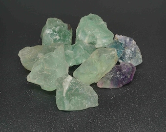 Puede incluir: Una colección de cristales de fluorita verde y púrpura. Los cristales son ásperos y tienen una variedad de formas y tamaños.