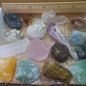May include: A collection of 15 different types of crystals and stones in a box. The crystals are arranged in a grid pattern and include amethyst, rose quartz, selenite, tiger's eye, and more.