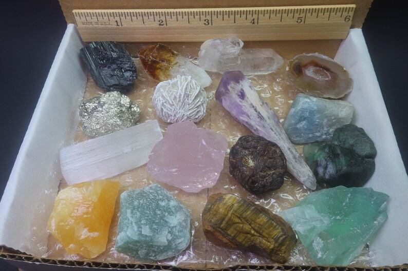 May include: A collection of 15 different types of crystals and stones in a box. The crystals are arranged in a grid pattern and include amethyst, rose quartz, selenite, tiger's eye, and more.