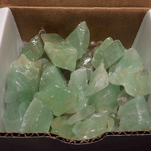 May include: A box filled with a large quantity of light green tumbled stones. The stones are smooth and have a translucent appearance.