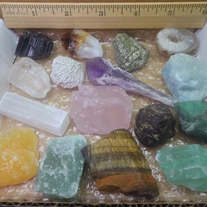 May include: A collection of 14 different types of crystals and stones in a white box. The crystals include amethyst, rose quartz, selenite, clear quartz, tiger's eye, pyrite, and more.