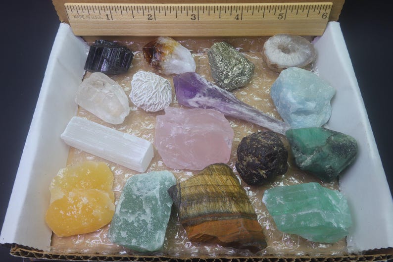 May include: A collection of 14 different types of crystals and stones in a white box. The crystals include amethyst, rose quartz, selenite, clear quartz, tiger's eye, pyrite, and more.