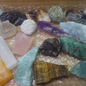 May include: A collection of 15 different types of crystals and stones, including amethyst, rose quartz, selenite, and tiger's eye. The crystals are arranged in a box with a brown background.