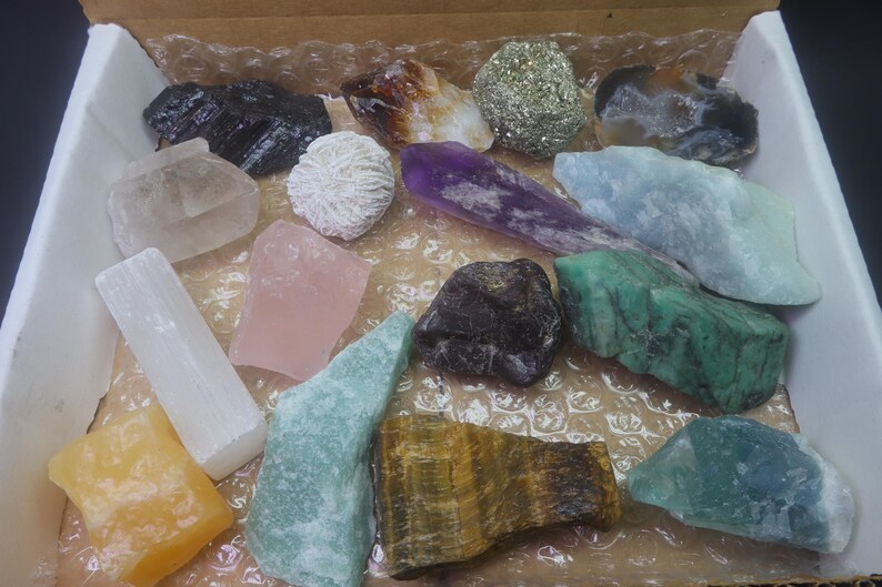 May include: A collection of 15 different types of crystals and stones, including amethyst, rose quartz, selenite, and tiger's eye. The crystals are arranged in a box with a brown background.
