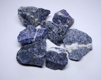 Puede incluir: Una colección de seis piedras de sodalita azul rugosas y naturales. Las piedras tienen una variedad de formas y tamaños, y algunas tienen vetas blancas que las atraviesan.