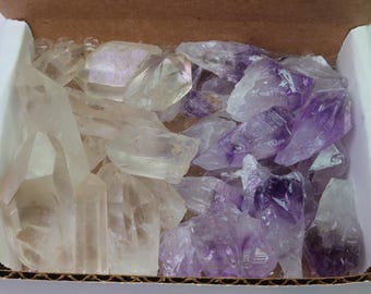 Puede incluir: Una caja de cartón llena de cristales en bruto sin cortar. Los cristales son una mezcla de cuarzo transparente y amatista, con los cristales de amatista que muestran tonos de púrpura. Los cristales son de varios tamaños y formas.