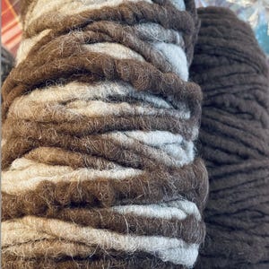 May include: Close-up of two yarn skeins. One skein has alternating bands of brown, light brown, and off-white. The other skein is a solid dark brown. The yarn appears thick and textured, suitable for knitting or crochet projects.