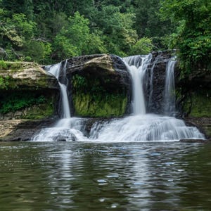 May include: A picturesque waterfall scene with water cascading over layered rocks into a tranquil pool. The rocks are covered in green vegetation, and the scene is framed by verdant trees.