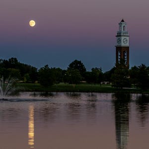 Puede incluir: Una serena escena crepuscular sobre un lago, con una luna llena en un cielo degradado púrpura y azul. Una fuente rocía agua y una torre del reloj se alza. El agua refleja la luna y la torre.
