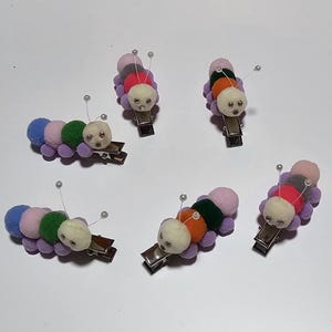 Pode incluir: Seis prendedores de cabelo em forma de lagarta coloridos com pompons e olhos saltados. As lagartas são feitas de feltro e têm diferentes combinações de cores.