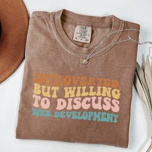 May include: A brown t-shirt with the text "Introverted, But Willing to Discuss Web Development" in colorful, retro-style lettering.