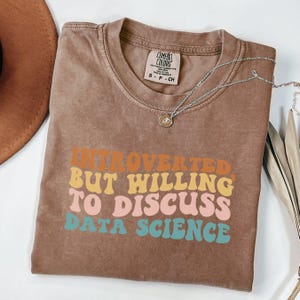 May include: A brown t-shirt with the text "INTROVERTED BUT WILLING TO DISCUSS DATA SCIENCE" in orange, yellow, pink, and teal. The shirt has a crew neck and short sleeves. A silver necklace with a pendant is draped across the shirt.
