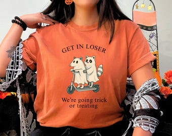 May include: Orange t-shirt with the text "GET IN LOSER" above an illustration of two ghost-covered animals on a scooter. Below the illustration is the text "We're going trick or treating."