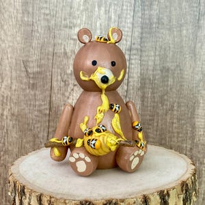 May include: A brown wooden bear figurine covered in honey and surrounded by small yellow and black bees. The bear is holding a small stick in its paws.