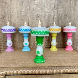 May include: Five colorful LED candle holders. Each has a different color base and ribbon trim, including purple, blue, green, yellow, and pink. The bases are decorated with flower accents. The candles have a flickering flame effect.