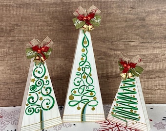 May include: Three wooden Christmas tree ornaments with green and gold painted designs. Each tree has a burlap bow with a red bell and a gold bell on top.