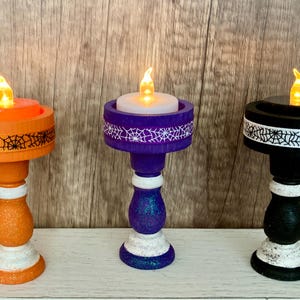 May include: Three Halloween-themed candle holders in orange, purple, and black. Each holder has a decorative spiderweb design and a flickering LED candle. The holders are painted with glitter accents and white trim.