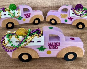 Puede incluir: Tres camiones de juguete de madera púrpura con camas blancas, decorados con cuentas doradas y verdes, monedas de oro y las palabras "Mardi Gras".