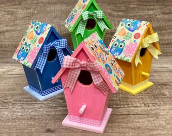 May include: Four colorful birdhouses with different patterned roofs. The birdhouses are painted in blue, green, pink, and yellow. Each birdhouse has a small opening for birds to enter and a perch for them to land on.