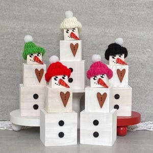 May include: Five wooden snowman ornaments stacked on top of each other. Each snowman has a different colored knitted hat, a carrot nose, and a heart-shaped design. The snowmen are on a white wooden base.