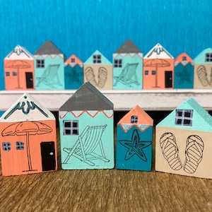 May include: A collection of ten miniature wooden houses painted in various shades of blue, green, and orange. Each house features a unique design, including a beach chair, a starfish, and flip-flops. The houses are arranged in a row on a white surface.