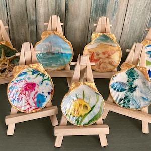 May include: Set of six seashell-shaped trinket dishes with various nautical scenes painted on them. Each dish is displayed on a small wooden easel.