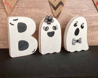 May include: White wooden ghost-shaped letters spelling "BOO" with black painted details. The "O" is a ghost with a bow tie and the "B" has bat silhouettes. The "O" has a black bow and eyelashes.