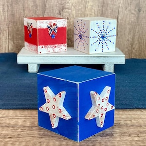 May include: Three wooden blocks painted in red, white, and blue with patriotic designs. The blocks feature a red bow, a white starburst, and two white stars with red polka dots.