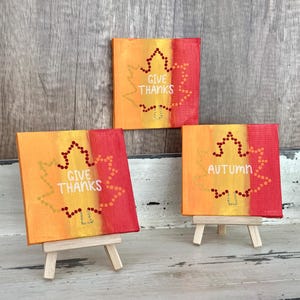 May include: Three small square canvases with autumn-themed art. Each canvas features a leaf design with the words "GIVE THANKS" or "AUTUMN" in white. The canvases are painted in orange and red hues and are displayed on small wooden easels.