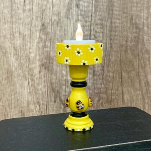 May include: A yellow candle holder with a floral design and a small LED candle. The candle holder has a round base and a decorative bee design. The top of the candle holder is yellow with white and black flowers.