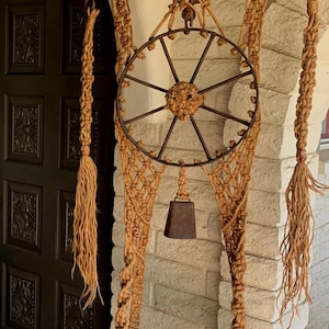 Può includere: Un portavasi in macramè con uno scaffale in legno e una ruota in metallo. Il portavasi è fatto di corda in fibra naturale e ha una campana appesa. Il portavasi è appeso a un muro di mattoni.