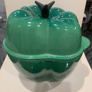 Könnte beinhalten: Ein grüner gusseiserner Topf in Form einer Paprika mit einem schwarzen Knopf auf dem Deckel.