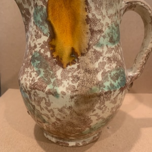 Può includere: Un'antica brocca in ceramica con una smaltatura marrone, bianca e verde screziata. La brocca ha un'ampia apertura e un manico.