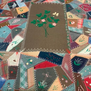 Może przedstawiać: Antyczna kołdra w patchworku z różnymi tkaninami i kolorami. Panel centralny ma brązowe tło z haftowanym wzorem kwiatowym. Kołdra składa się z wielu różnych kształtów i rozmiarów kawałków tkaniny, w tym kwadratów, trójkątów i prostokątów. Kołdra jest zszyta białą nicią.